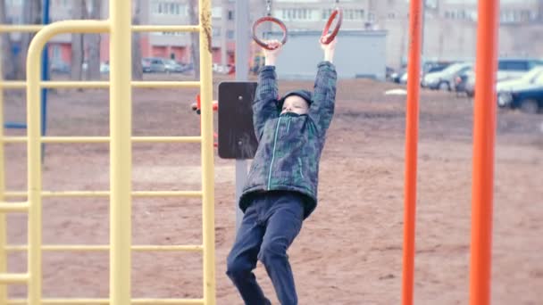 Jongen swingt op gymnastiek ringen, sprongen en glimlacht. Speeltuin in de binnenplaats van het city house. — Stockvideo