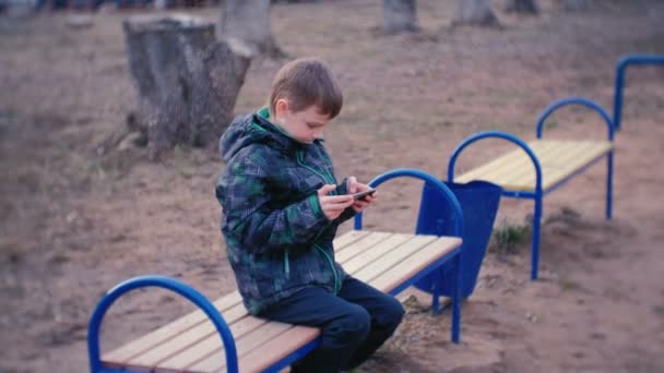 Menino joga um jogo em seu telefone celular sentado no parque em um banco . — Vídeo de Stock