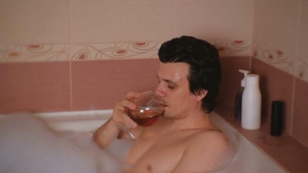 Young man takes a bath with foam and drinks whiskey. Smiling and looking at camera. — Stock Video