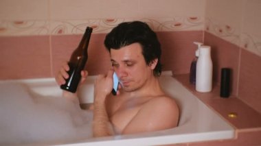 Genç adam bir banyo köpüğü ile alır, cep telefonundan konuşuyor, bira şişesinden içiyor