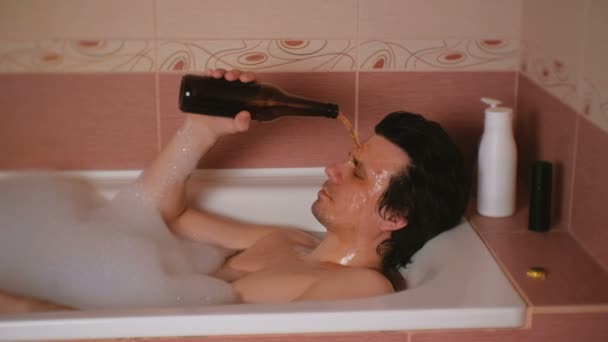 Ubriaco uomo versa birra da una bottiglia sul suo viso sdraiato in bagno con schiuma . — Video Stock