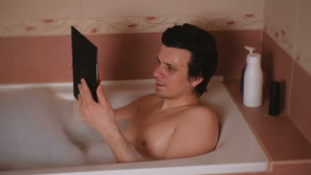 Молодой человек принимает ванну, просматривает интернет на планшете и смотрит в камеру . — стоковое видео