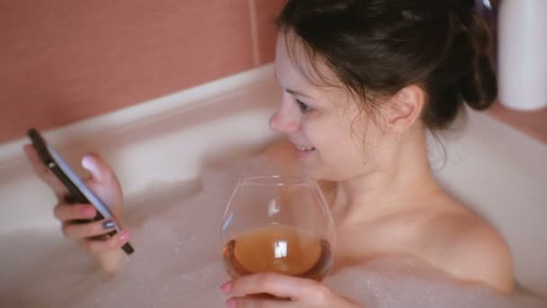 Giovane bella donna si fa un bagno, navigando in internet su un telefono cellulare e beve whisky . — Video Stock