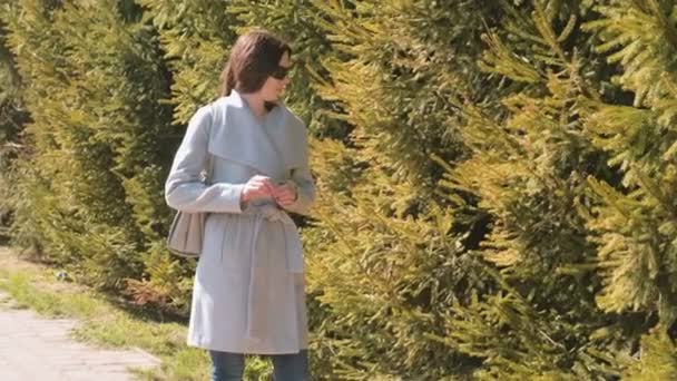 Mladá žena v kabát čeká na někoho v městském parku a sluneční brýle. Jarní čas. — Stock video