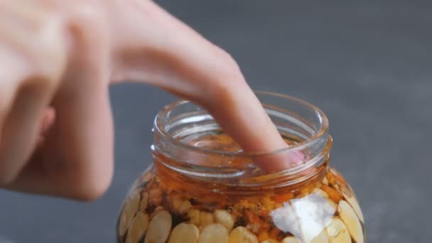 Woman dips her finger in honey with nuts in the jar. Close-up hand, finger and jar on black table. — Stock Video