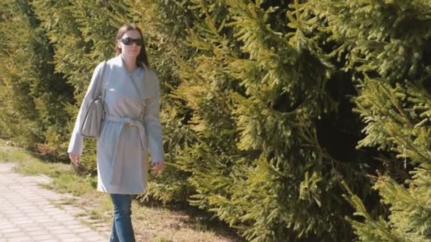 Unga brunett vacker kvinna i solglasögon och coat promenader i stadsparken i soliga vårdag. — Stockvideo