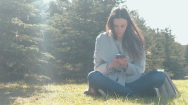 Genç güzel esmer kadın bahar Park çimenlerin üzerinde oturur ve hareket eden telefon içinde bir ileti türleri.