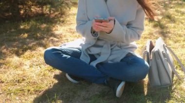 Genç güzel esmer kadın bahar Park çimenlerin üzerinde oturur ve hareket eden telefon içinde bir ileti türleri. Yakın çekim yakışıklı.