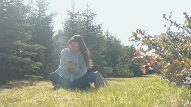 Jonge mooie brunette vrouw zit op gras in voorjaar park op wind en een bericht typen in mobiele telefoon. — Stockvideo