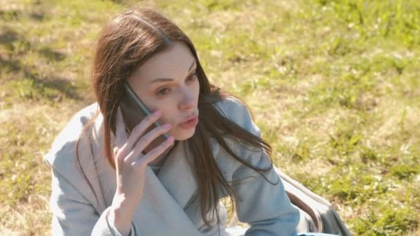 Portrait of young beautiful woman sitting in the spring park on the grass and talking on a mobile phone. — Stock Video