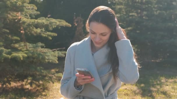 Jonge mooie brunette vrouw zit op gras in voorjaar park en een bericht typen in mobiele telefoon, camera en lachend kijkt. — Stockvideo