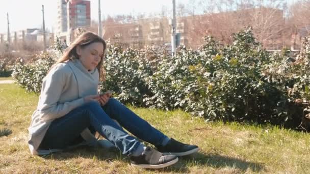 Joven mujer morena hermosa se sienta en la hierba en el parque de primavera, escuchar música en el teléfono y cantar una canción . — Vídeo de stock