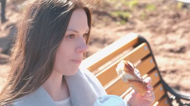Die junge brünette Frau im Mantel sitzt auf einer Bank im Stadtpark, isst Brötchen mit Zucker und trinkt Kaffee. Frühling. Gesicht aus nächster Nähe. — Stockvideo
