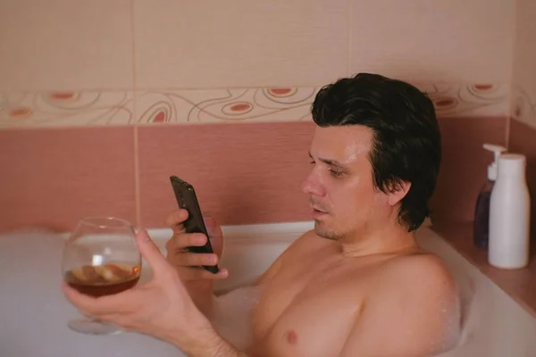 Jonge man neemt een bad met schuim, typen bericht op mobiele telefoon en dranken-whisky en glimlach. — Stockfoto
