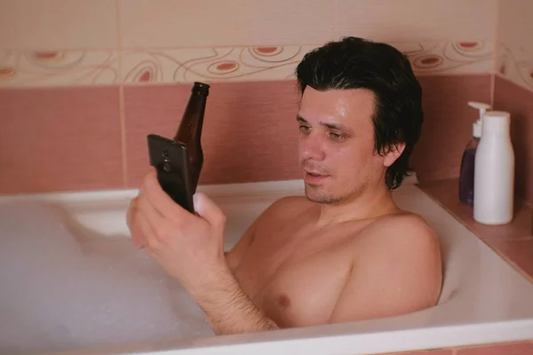 Молодой человек принимает ванну с пеной, просматривает интернет на мобильном телефоне, пьет пиво из бутылки . — стоковое фото