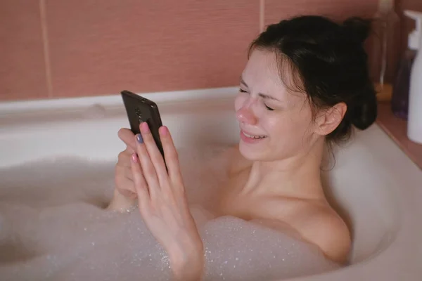 Jonge mooie brunette vrouw neemt een bad, leest een bericht op mobiele telefoon en kreten. — Stockfoto