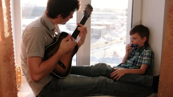 楽器を演奏します パパがギターを弾いているし 息子が窓辺に座って口臓器を再生 — ストック動画