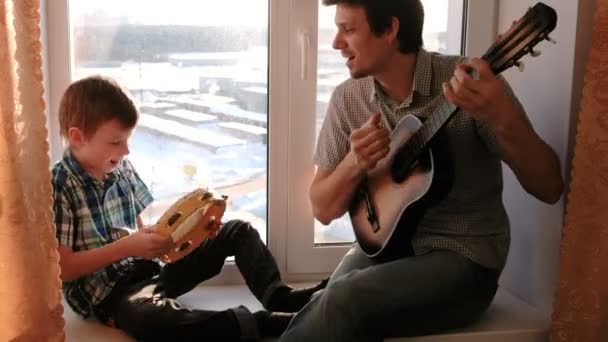 Грає на музичному інструменті. Тато грає на гітарі, а син грає на тамбурі, сидячи в підвіконні . — стокове відео
