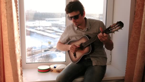 Het spelen van een muziekinstrument. Man speelt de gitaar zitten in de vensterbank. — Stockvideo