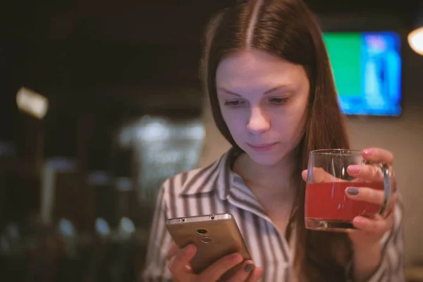 Ung, vakker brunette leser noe på mobilen og drikker rød urtete på kafeen. . – stockfoto