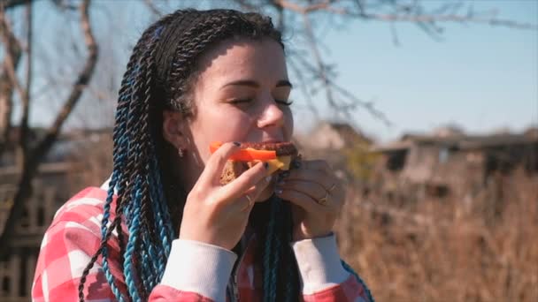 Junge Frau mit blauen Flechthaaren isst Sandwich mit Brot, Schnitzel, Pfeffer und Käse im Freien. — Stockvideo