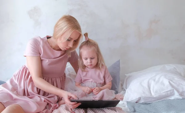 Mamma och dotter titta i tablett. Ung attraktiv blond kvinna med hennes lilla charmiga dotter i rosa klänningar du tittar på något i tablett. — Stockfoto