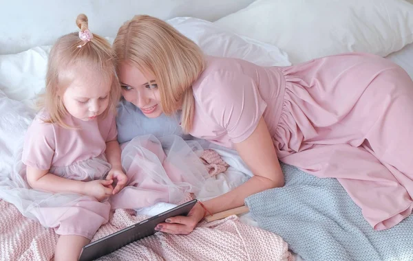 Молодая привлекательная блондинка учит свою маленькую очаровательную дочь в розовых платьях, используя планшет, лежащий на кровати . — стоковое фото