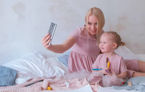 Молодая привлекательная блондинка мама показывает что-то в мобильном телефоне своей маленькой очаровательной дочери в розовых платьях. Или делает селфи — стоковое фото