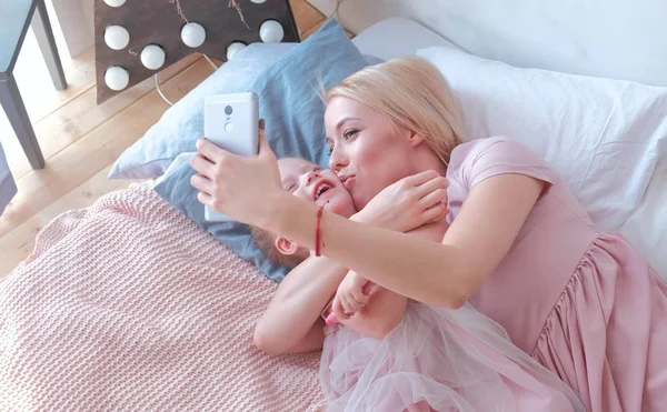 Young attractive blond mom shows something in mobile phone to her little charming daughter in pink dresses. Or makes selphie