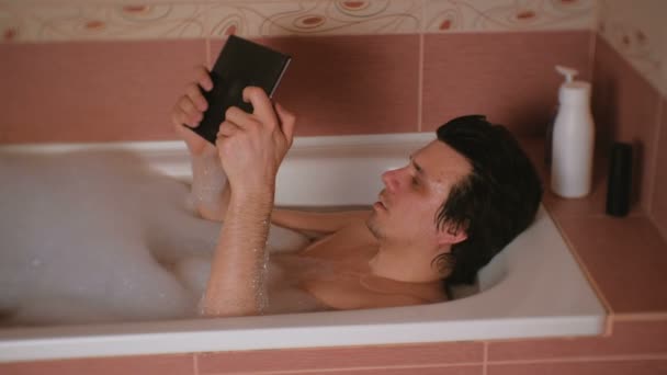 Młody człowiek bierze kąpiel, gra w gry na tablet. — Wideo stockowe