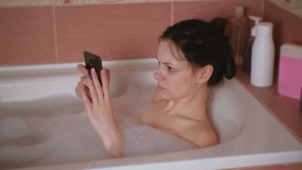 Junge schöne brünette Frau badet, schaut ein Video auf dem Handy. — Stockvideo