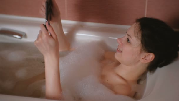 Молода красива брюнетка приймає ванну і дивиться смішне відео на планшет . — стокове відео