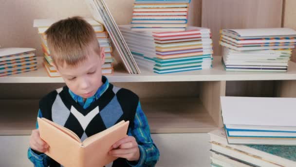 Rapaz lê um livro sentado no chão entre os livros . — Vídeo de Stock