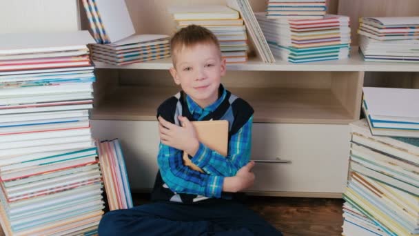 Menino de sete anos abraçando o livro e sorrindo para a câmera sentada entre os livros. Fechar. . — Vídeo de Stock