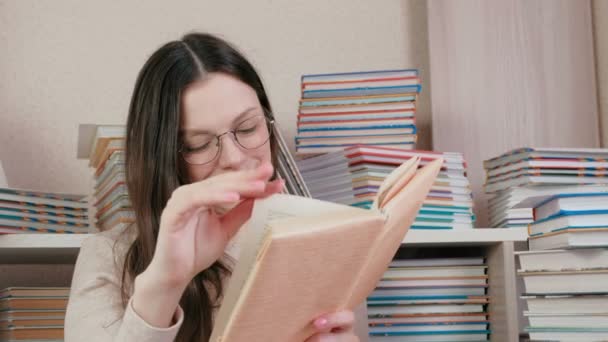 Ung brunett kvinna i glasögon leende en bok. Sitter bland böckerna. — Stockvideo