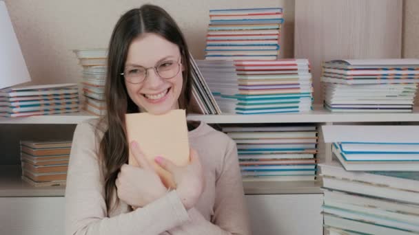 Młoda kobieta brunetka w okularach śmieje się, przytulanie książki siedzi wśród książek. — Wideo stockowe