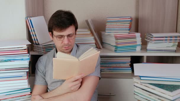 Junger brünetter Mann mit Brille liest ein Buch sitzend zwischen Büchern.