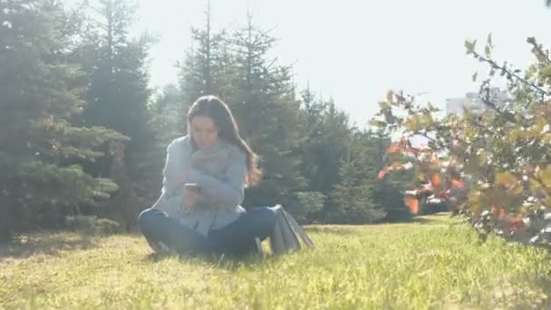 Jonge mooie brunette vrouw zit op gras op sterke wind in voorjaar park en een bericht typen in mobiele telefoon. — Stockvideo