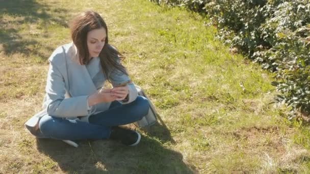 Jovem bela mulher sentada no parque da primavera na grama e falando em um telefone celular . — Vídeo de Stock