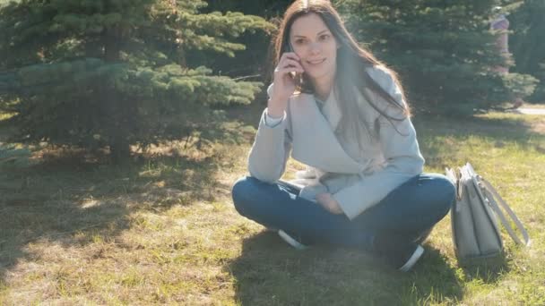 Retrato de la joven feliz hermosa mujer sentada en el parque de primavera en la hierba y hablando en un teléfono móvil . — Vídeo de stock