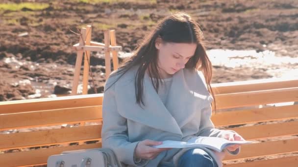 Young beautiful brunette woman student sits on the bench in spring park, reads a book — Stock Video