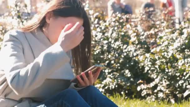 Jovem bela mulher morena senta-se na grama no parque da primavera, olha as mídias sociais no telefone móvel . — Vídeo de Stock