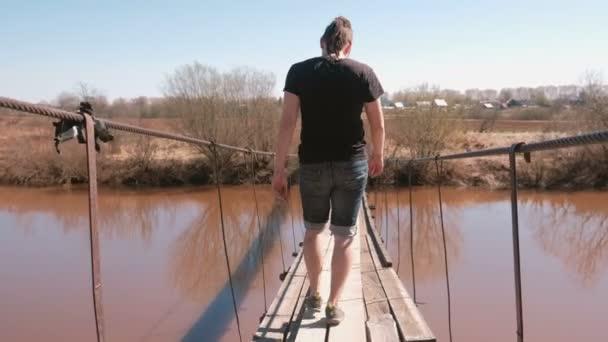 Молодой человек идет по взвешенному деревянному мосту через реку. Вид сзади . — стоковое видео
