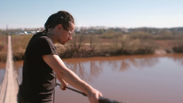 Un joven está parado en un puente de madera suspendido sobre el río. . — Vídeos de Stock