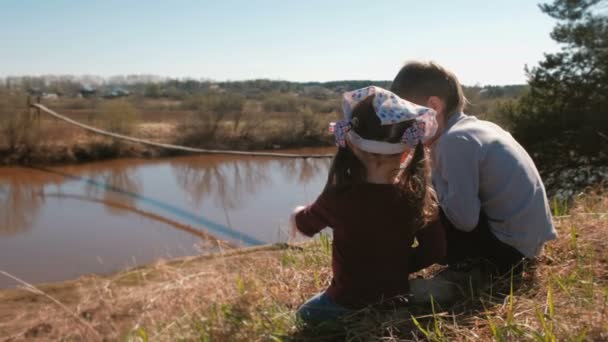 Hermano y hermana sentados y jugando en la orilla del río en el pueblo. Vista trasera . — Vídeos de Stock