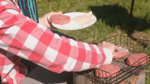 Une jeune femme met des escalopes sur le gril. Gros plan sur les mains. Elle est en chemise rose à carreaux et aux cheveux tressés bleus . — Video