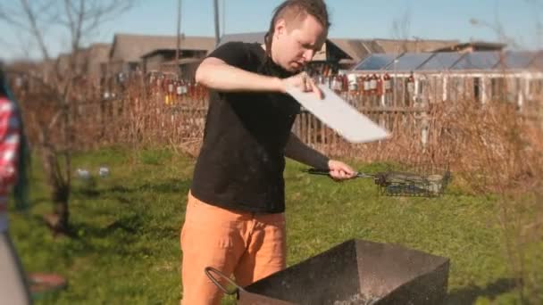 Молодой человек надувает угли в гриле жаровней с помощью разделочной доски . — стоковое видео