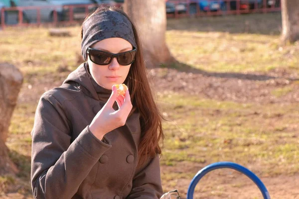 Молодая брюнетка в солнечных очках ест мандарин, сидя на скамейке в парке. . — стоковое фото