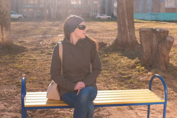 Joven hermosa morena en gafas de sol mira a su alrededor y espera a alguien sentado en el banco en el patio de la ciudad de varios pisos . — Foto de Stock