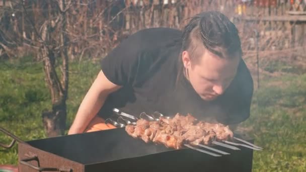 Young man with dreads on hairs cooks shashlik meat on top of charcoal grill on backyard. — Stock Video